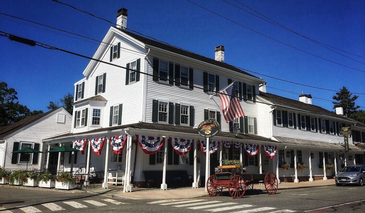 The historic Griswold Inn in Essex, Connecticut, adorned with American flags and patriotic bunting. The white building with black shutters and a covered porch exudes classic New England charm under a bright blue sky. An antique wagon is displayed out front, enhancing the inn's nostalgic atmosphere.