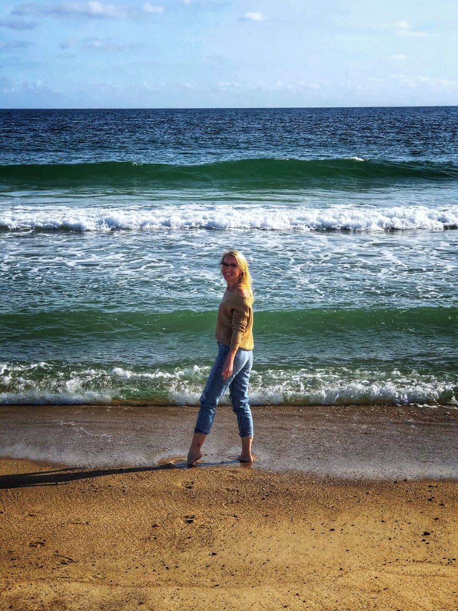 Kate walks barefoot along a sandy beach in Rhode Island, with gentle waves lapping at the shore. She is wearing a light sweater and rolled-up jeans, enjoying the serene ocean view under a partly cloudy sky.