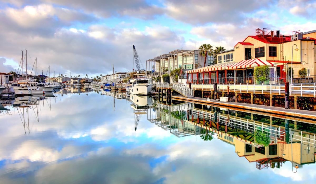 A tranquil harbor scene in Newport, Rhode Island, with boats docked along the calm water reflecting the sky and surrounding buildings. A red and white striped canopy adorns a waterfront building, adding a pop of color to the picturesque setting. The sky is partly cloudy, creating a serene and inviting atmosphere.