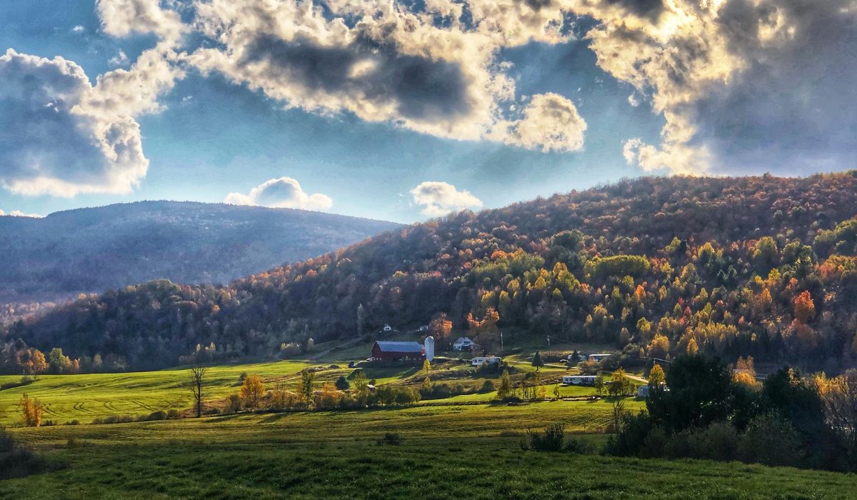 A picturesque Vermont valley in the fall, featuring a red barn and silos nestled among rolling green fields. The surrounding hills are covered in trees with vibrant autumn foliage under a dramatic sky with scattered clouds. The scene captures the serene beauty of rural Vermont during the fall season.