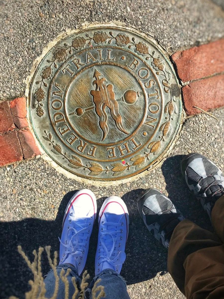 A close-up view of two people standing next to a bronze marker on the Freedom Trail in Boston. The marker features an engraved arrow and the text "The Freedom Trail Boston," surrounded by a decorative border. 