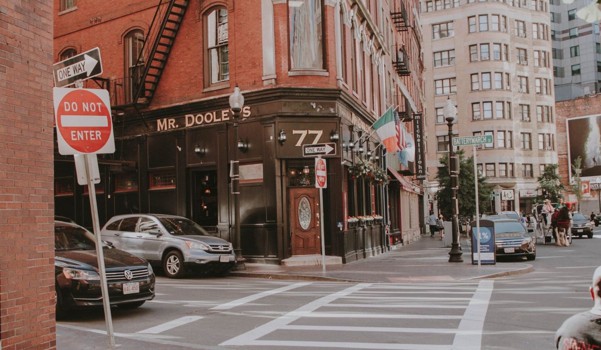 A street view in Boston, Massachusetts, featuring the corner pub "Mr. Dooley's" at 77 Batterymarch Street. The pub has a traditional black and brick facade with an Irish flag hanging outside.