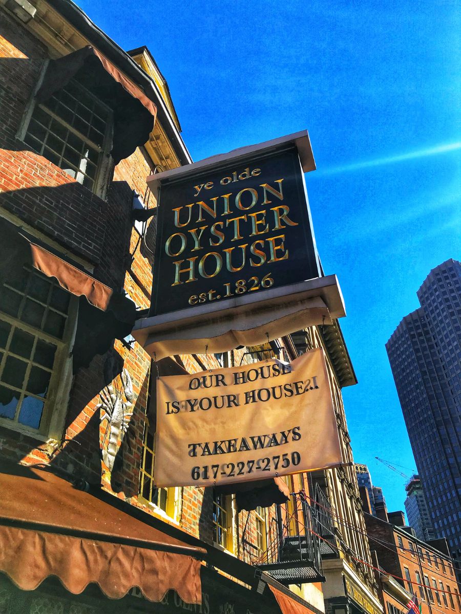 The exterior of Ye Olde Union Oyster House in Boston, established in 1826, featuring a large sign that reads "Our House Is Your House!"  The historic brick building is bathed in sunlight against a clear blue sky, with modern skyscrapers visible in the background.