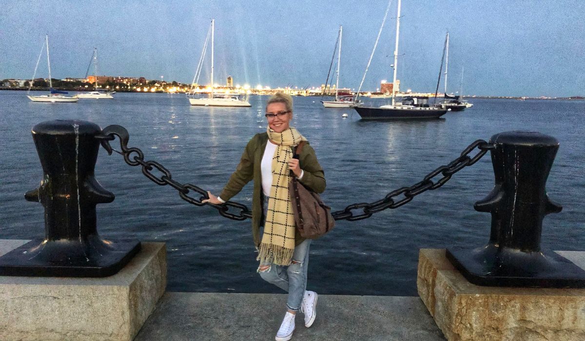 Kate stands by the waterfront in Boston, leaning on a large black chain link fence with several sailboats anchored in the harbor behind her. She is wearing a green jacket, a beige plaid scarf, jeans, and white sneakers, with city lights twinkling in the distance as evening falls