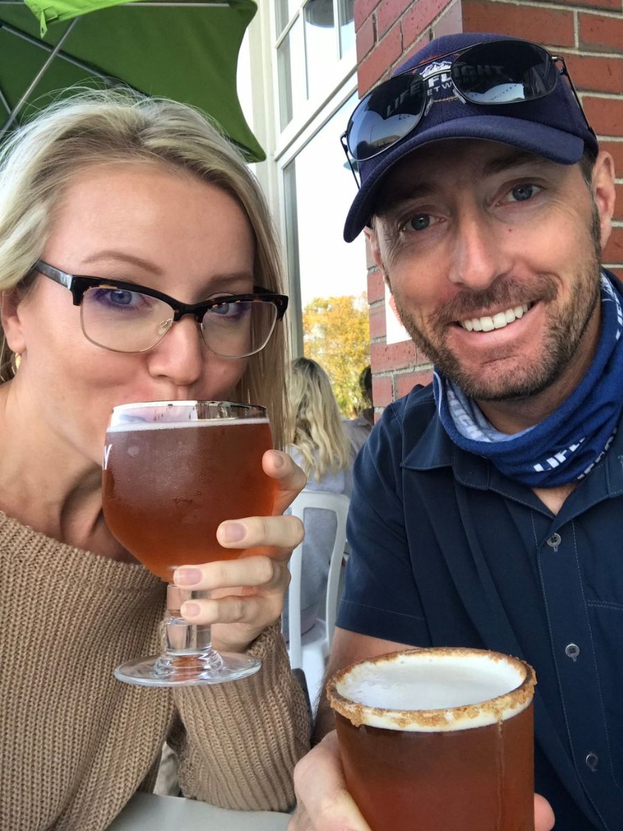 Kate and her husband enjoy pumpkin beer at an outdoor café in Newburyport, Mass. Kate is wearing glasses and a beige sweater, while her husband sports a blue cap and shirt, smiling at the camera.