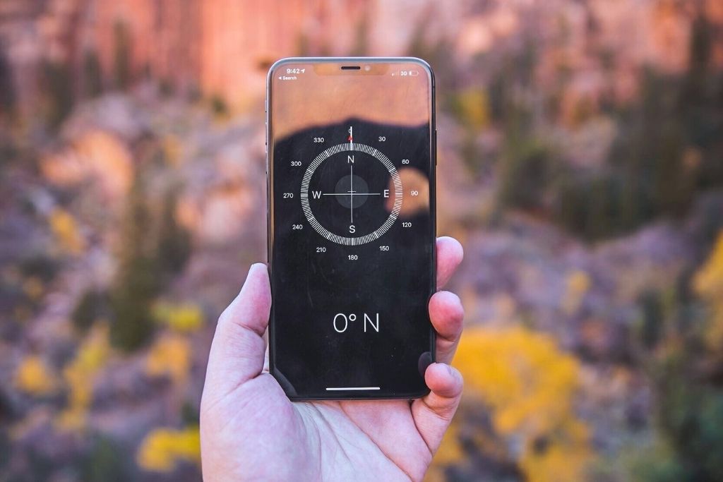 This image shows a hand holding a smartphone displaying a digital compass reading "0° N," with a blurred natural background featuring warm tones of yellow and orange foliage.