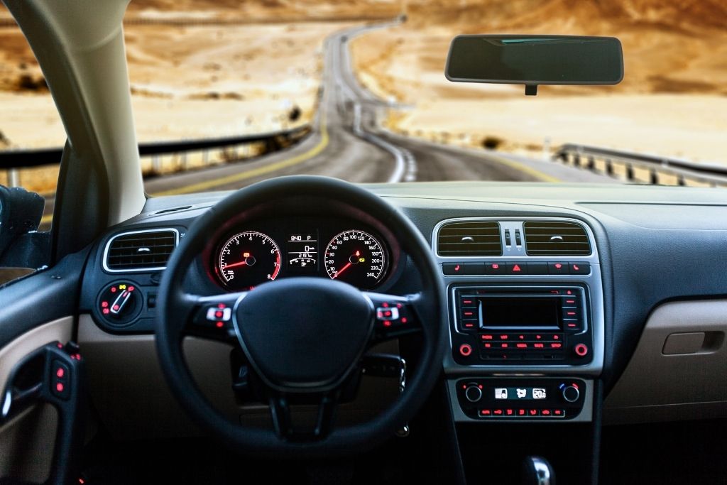 This image shows the interior view of a car, focusing on the dashboard and steering wheel, with a winding desert road visible through the windshield. The setup suggests a road trip adventure, highlighting the journey through an arid, sandy landscape.