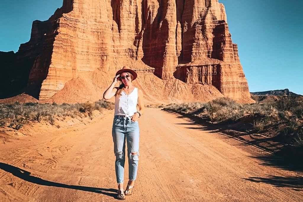 Kate wearing a pair of blue jeans, a white tank top, and a brown hat. She is standing in the middle of a dirt road in Utah with red rock spires standing behind her with a clear blue sky.