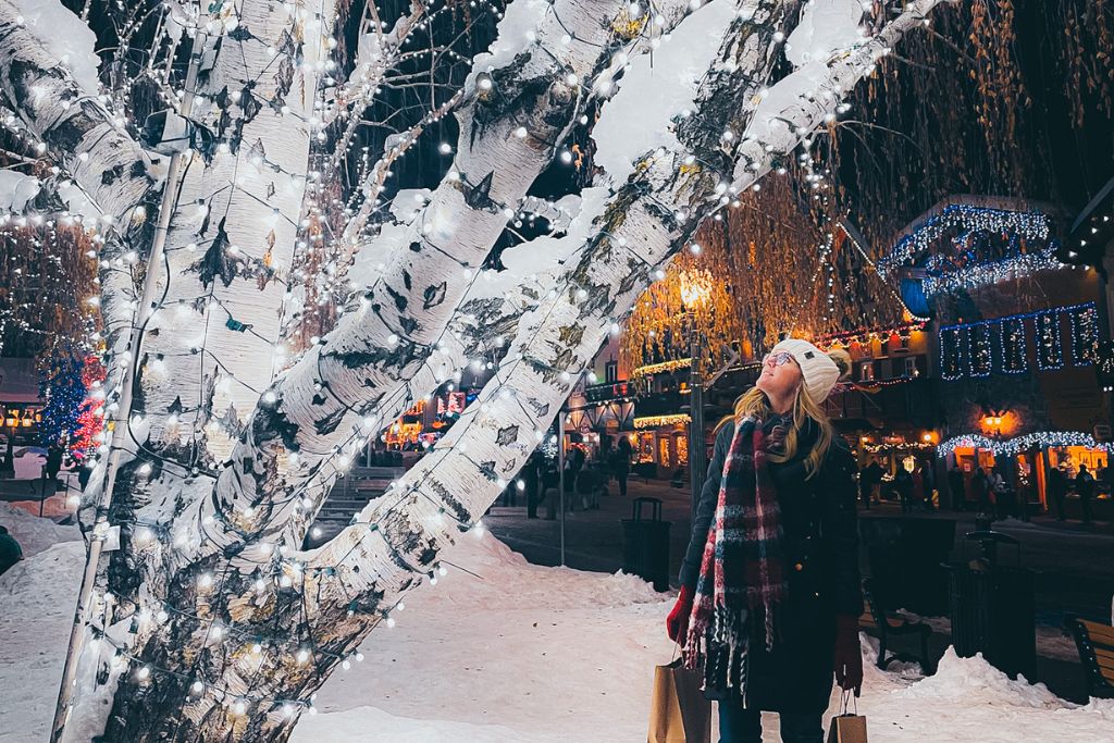 Kate from Kate's Crossing Blog is dressed warmly in a black coat, plaid scarf, and white beanie looks up in awe at a snow-covered birch tree wrapped in twinkling white lights. The scene is set in Leavenworth, Washington, where the festive Christmas lights glow on the Bavarian-style buildings in the background. The snow-covered ground and decorated tree create a magical "Christmas in Leavenworth" atmosphere. She holds shopping bags, enjoying the holiday season.