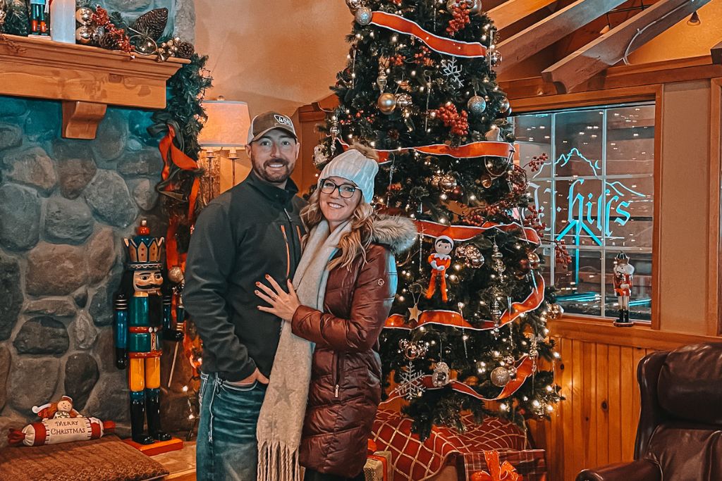 Kate from Kate's Crossing Blog and her husband stand in front of a beautifully decorated Christmas tree inside a cozy lodge, embracing and smiling for the camera. Kate is bundled up in a winter coat and beanie, while her husband wears a baseball cap and jacket. The room is decorated with festive holiday decorations, including a large nutcracker and a stone fireplace. The glowing tree is wrapped with red ribbon and ornaments, creating a warm, festive atmosphere perfect for celebrating Christmas in Leavenworth, Washington.