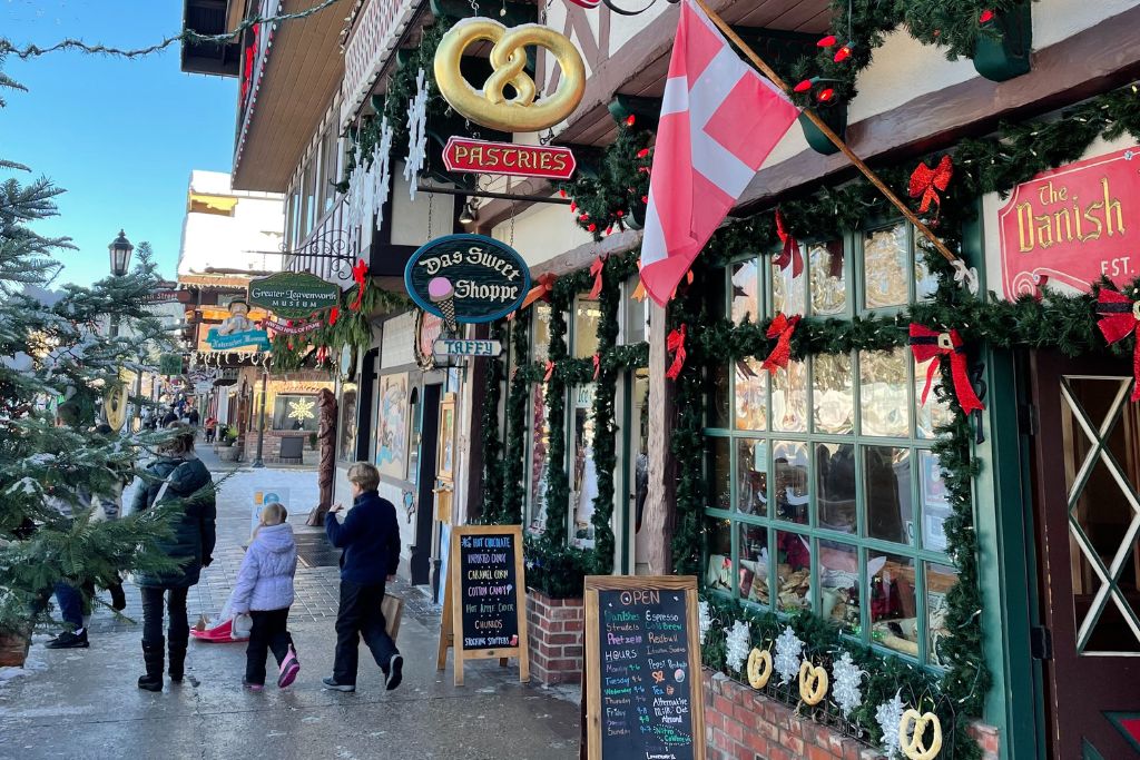 A charming street in Leavenworth, Washington, lined with Bavarian-themed shops decorated for the holiday season. The storefronts feature garlands, red bows, and festive signs, including a large pretzel symbol for a pastry shop. A Danish flag hangs outside "The Danish" bakery, while people walk along the sidewalk, bundled up in winter coats. 