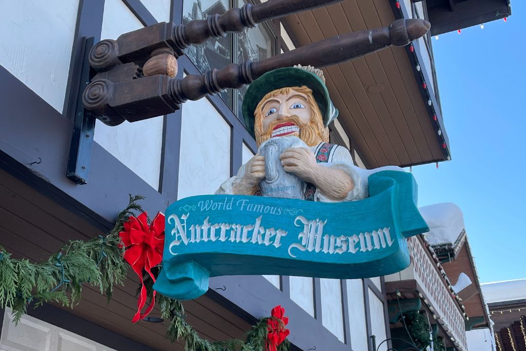 A colorful sign for the "World Famous Nutcracker Museum" hangs outside a Bavarian-style building in Leavenworth, Washington. The sign features a large carved figure of a bearded man wearing traditional Bavarian clothing and holding a beer stein, resembling a nutcracker. Festive garlands and red bows decorate the building, adding to the holiday spirit in this charming Christmas town.