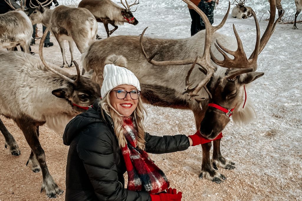 Kate from Kate's Crossing Blog is dressed in a winter coat, plaid scarf, and white beanie kneeling next to a group of reindeer in a snowy setting in Leavenworth, Washington. She smiles while petting one of the reindeer, which has large antlers and is wearing a red harness. 