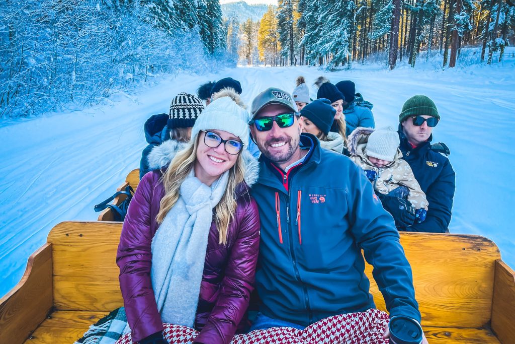 Kate from Kate's Crossing and her husband smile while sitting bundled up in blankets on a wooden sleigh during a winter sleigh ride in Leavenworth, Washington. They are surrounded by other riders, all enjoying the snowy landscape of frosted trees and a snow-covered trail. The bright winter day and serene scenery create a joyful and cozy holiday experience in this festive setting.