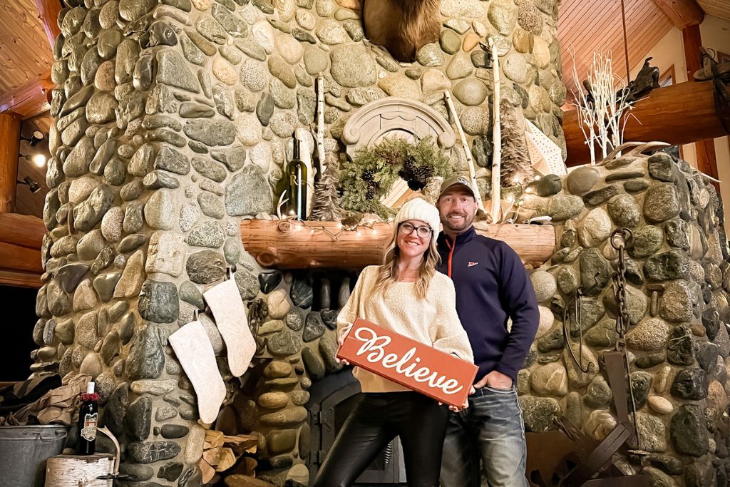 Kate and her husband stand in front of a large stone fireplace inside a cozy cabin, smiling and holding a sign that reads "Believe." The mantle is decorated with stockings, wreaths, and holiday greenery, creating a festive Christmas atmosphere. The warm, rustic setting, complete with wood beams and a mounted animal head above the fireplace