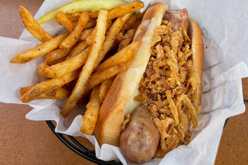 A basket of food featuring a grilled sausage topped with crispy fried onions in a toasted bun, served alongside seasoned French fries and a pickle spear. The meal is presented in a paper-lined basket, offering a hearty and satisfying dish, likely enjoyed at a local eatery in Leavenworth, Washington, during the holiday season.