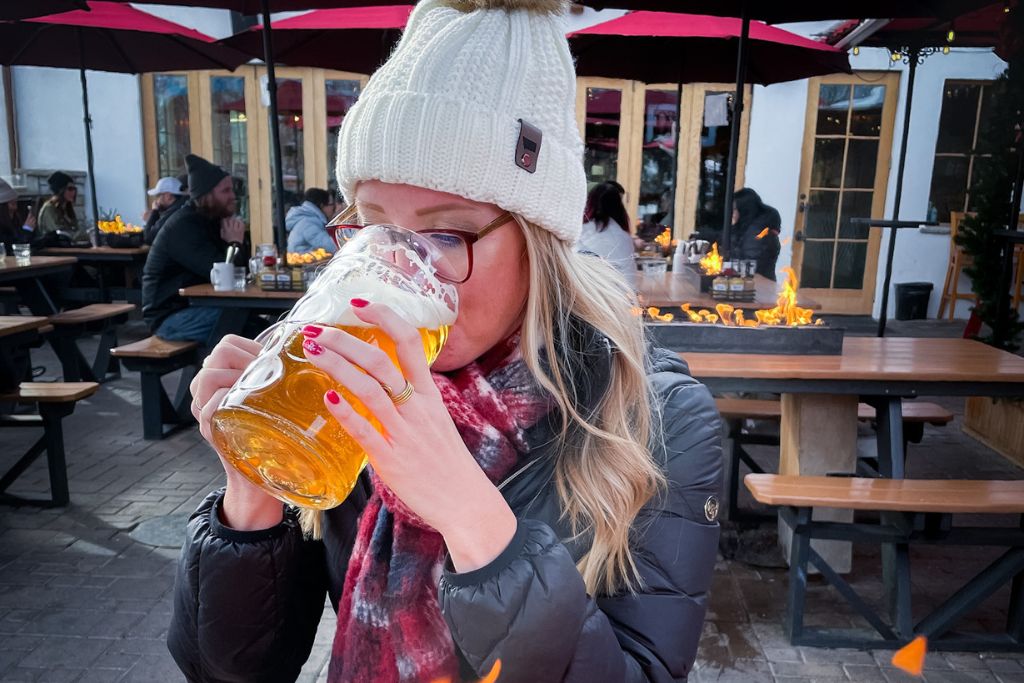 Kate of Kate's Crossing Blog in a winter beanie and jacket sips from a large glass of beer at an outdoor beer garden in Leavenworth, Washington. She is seated at a wooden picnic table near a fire pit, with other patrons enjoying the cozy atmosphere of the outdoor space. The warm ambiance and festive winter attire suggest a relaxing moment during the holiday season.