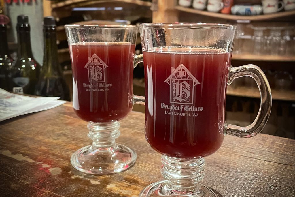 Two clear glass mugs filled with warm mulled wine sit on a wooden bar top at Bergdorf Cellars in Leavenworth, Washington. The mugs feature the winery’s logo, adding a festive touch to the cozy atmosphere. The deep red color of the wine and the inviting ambiance suggest a perfect way to enjoy the holiday season in this Bavarian-themed town.