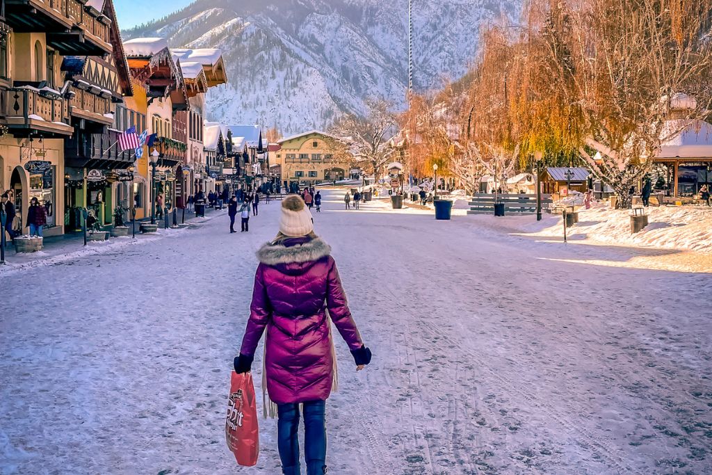 Kate of Kate's Cossing Blog is dressed in a red winter coat, and a white beanie walks down a snow-covered street in Leavenworth, Washington, carrying a shopping bag. The Bavarian-style buildings lining the street are adorned with festive decorations, while the surrounding snow-covered mountains create a picturesque winter backdrop.