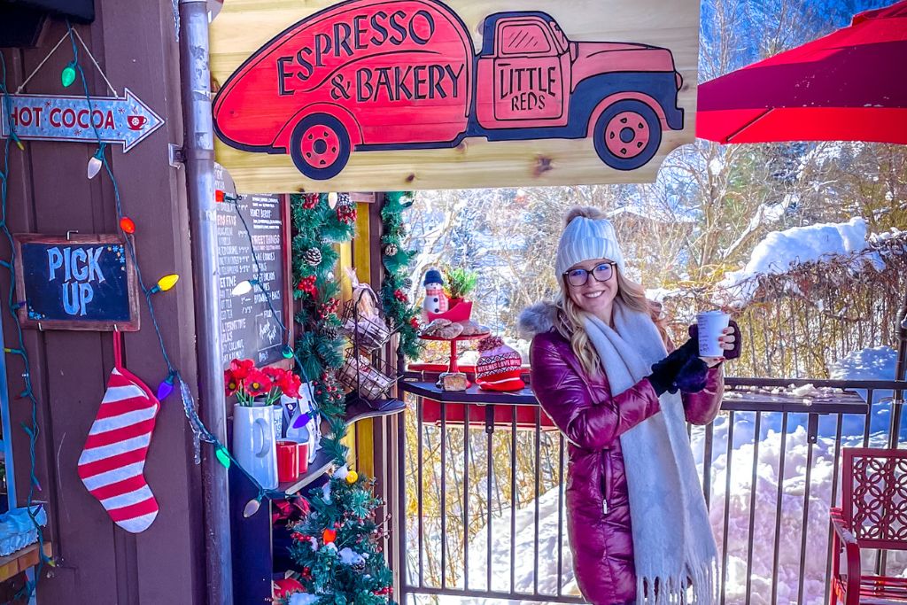 Kate in a red winter coat and white beanie holds a cup of coffee outside Little Reds Espresso & Bakery in Leavenworth, Washington. The cozy shop is decorated with festive garlands, a snowman figurine, and colorful holiday lights. A wooden sign featuring a red truck promotes the bakery, while a nearby arrow points to "Hot Cocoa" and "Pick Up." The snow-covered landscape adds to the festive, wintery atmosphere.