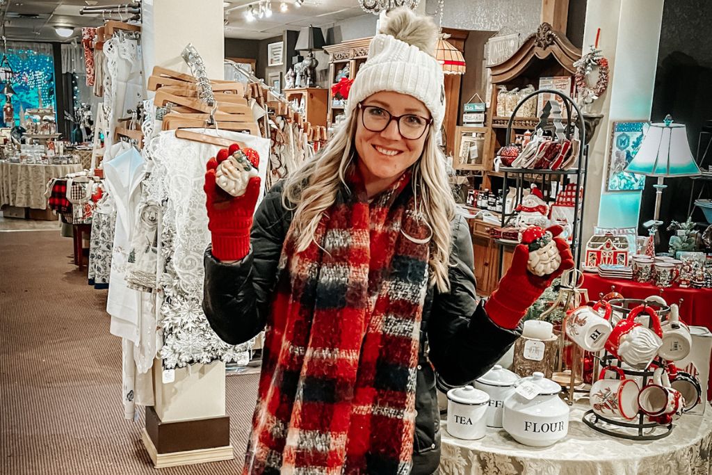 Kate of Kate's Crossing Blog is in a winter coat, plaid scarf, and white beanie smiles while holding two festive ornaments in a holiday shop in Leavenworth, Washington. The store is filled with Christmas decorations, kitchenware, and seasonal gifts, creating a cozy and inviting atmosphere. Kates red gloves and colorful ornaments add to the festive charm of the shopping experience during the holiday season.