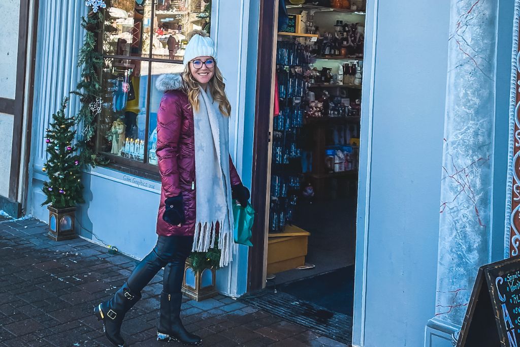 Kate in a red winter coat, white beanie, and scarf stands smiling outside a shop in Leavenworth, Washington. She holds a green shopping bag, having just stepped out of the store, which is decorated with festive garlands and a small Christmas tree by the window.