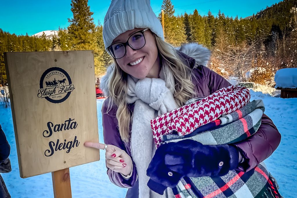 Kate's Crossing blog, stands in a snowy landscape holding cozy blankets while pointing to a wooden sign that reads "Santa Sleigh." She is bundled up in a white beanie, scarf, and a winter coat, ready for a sleigh ride in Leavenworth, Washington. The backdrop features snow-covered trees and mountains, capturing the festive and wintery spirit of the area.