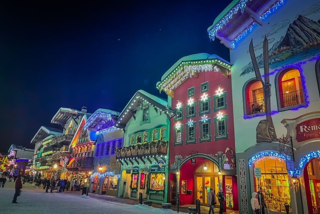 A vibrant nighttime view of the Bavarian-style buildings in downtown Leavenworth, Washington, beautifully adorned with colorful Christmas lights. The buildings, painted in festive hues of red, green, and white, feature intricate decorations, glowing snowflakes, and icicle lights. People stroll through the snowy street, enjoying the magical holiday atmosphere under a clear dark sky.