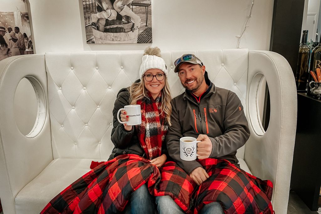 Kate from Kate's Crossing Blog and her husband sit cozily on a tufted white bench, wrapped in a red and black plaid blanket while holding mugs of hot mulled wine. Kate wears a white beanie and plaid scarf, while her husband sports a baseball cap and outdoor jacket. Behind them are black-and-white photos, creating a relaxed and warm atmosphere, perfect for enjoying a winter day in Leavenworth, Washington.