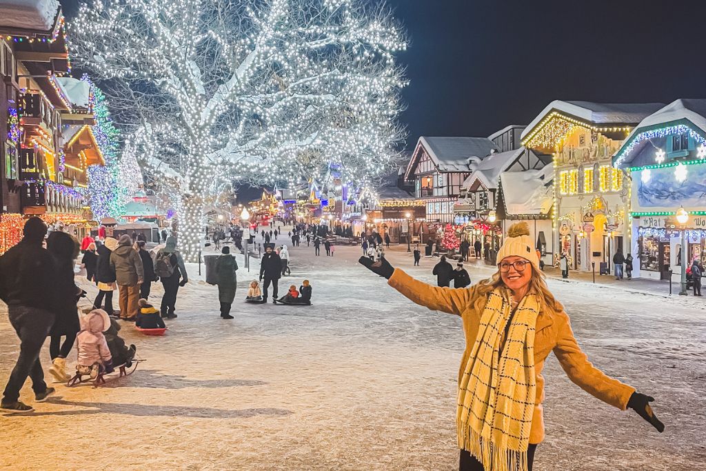 A festive winter scene in Leavenworth, Washington, with streets covered in snow and lined with Bavarian-style buildings illuminated by colorful Christmas lights. In the foreground, a smiling Kate from Kate's Crossing Blog is in a tan coat and scarf, wearing a yellow beanie, poses with arms outstretched. Families and children can be seen enjoying the holiday atmosphere, some sledding down the snowy street. The trees are wrapped in bright white lights, adding to the magical "Christmas in Leavenworth" feel.