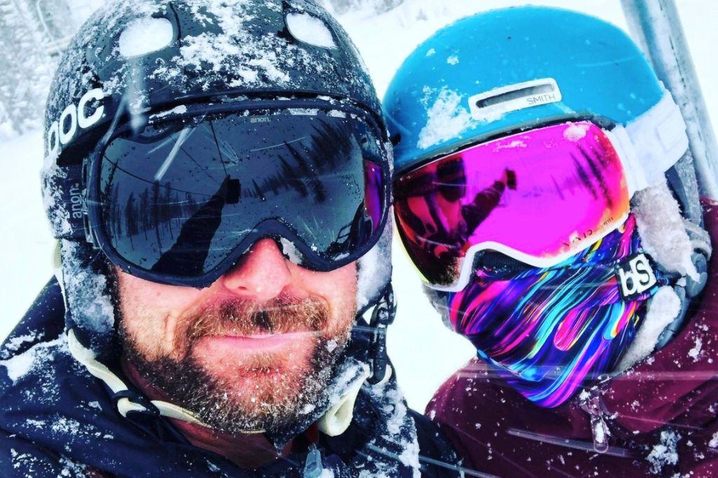 Kate and her partner are wearing helmets, goggles, and face coverings, enjoying a snowy ski day. Their gear is dusted with snow, highlighting the cold and adventurous conditions of the slopes.