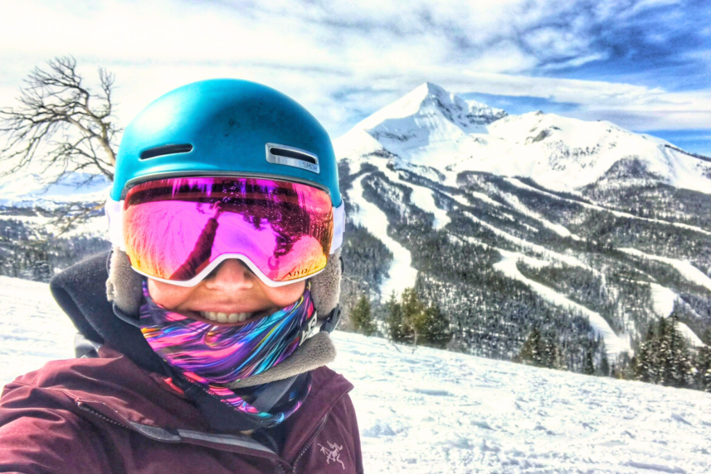 Kate taking a selfie on a snowy slope, wearing a vibrant blue helmet and reflective goggles that reflect the surrounding landscape. The backdrop showcases a majestic snow-covered peak and ski runs under a bright blue sky