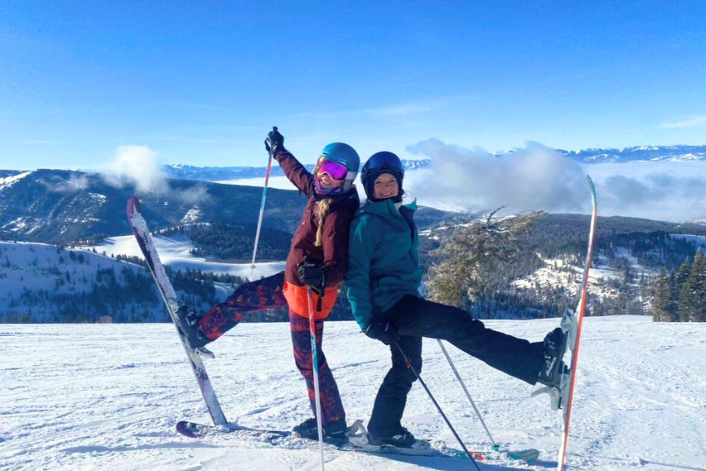 Two skiers posing playfully on a snowy mountaintop, each balancing on one leg with their skis and poles. The bright blue sky and expansive snow-covered mountain landscape in the background create a vibrant and joyful winter scene.