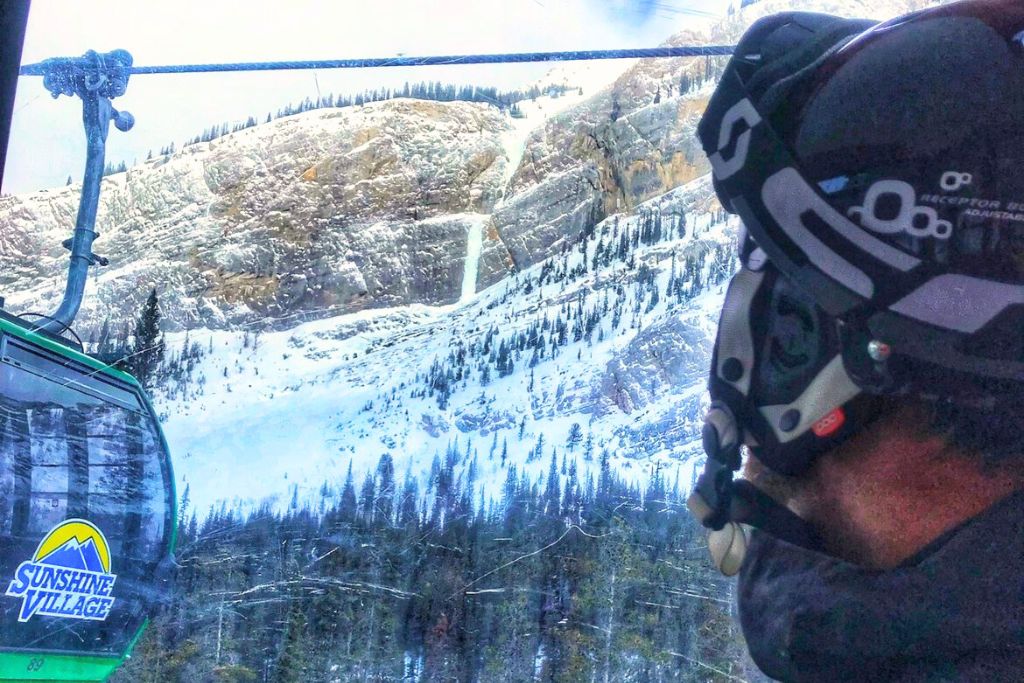 This image features a skier riding a gondola at Sunshine Village, gazing at a rugged mountain covered in snow and rock formations. The gondola window frames a stunning view of the alpine scenery.