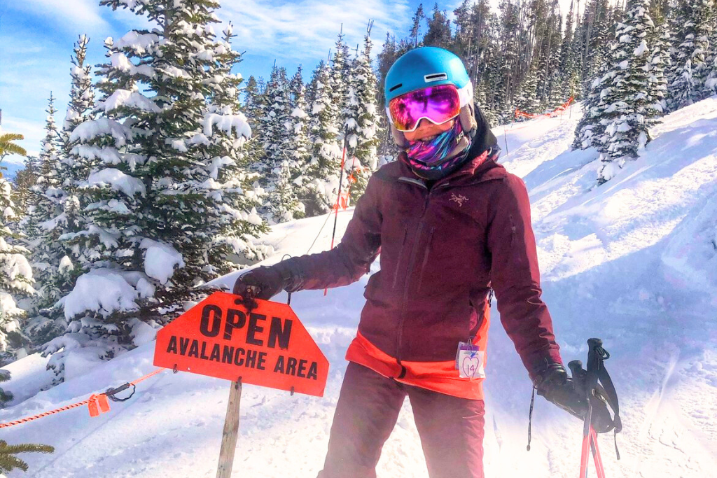 Kate posing next to a bright orange sign that reads "Open Avalanche Area," surrounded by snow-covered trees on a sunny day. She is wearing colorful, reflective goggles and a blue helmet, with a backdrop of pristine snow and a vibrant blue sky.