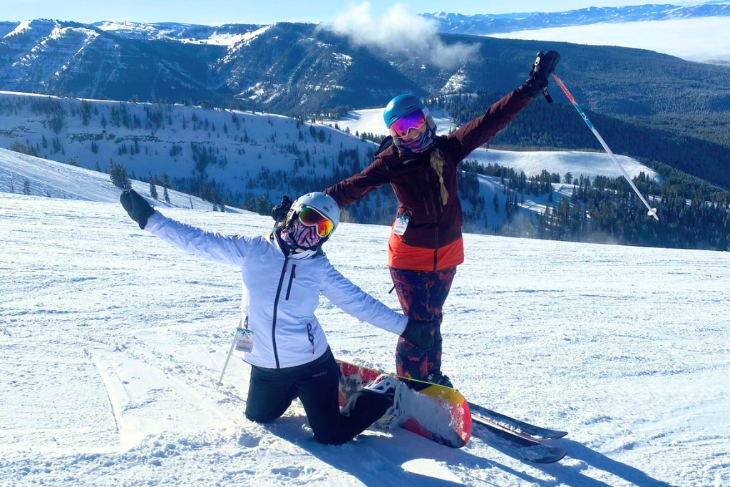 Kate and her friend posing on a snowy slope with wide smiles and arms outstretched, showcasing their skis and snowboard. The backdrop features a bright blue sky and sweeping views of snow-covered mountains.