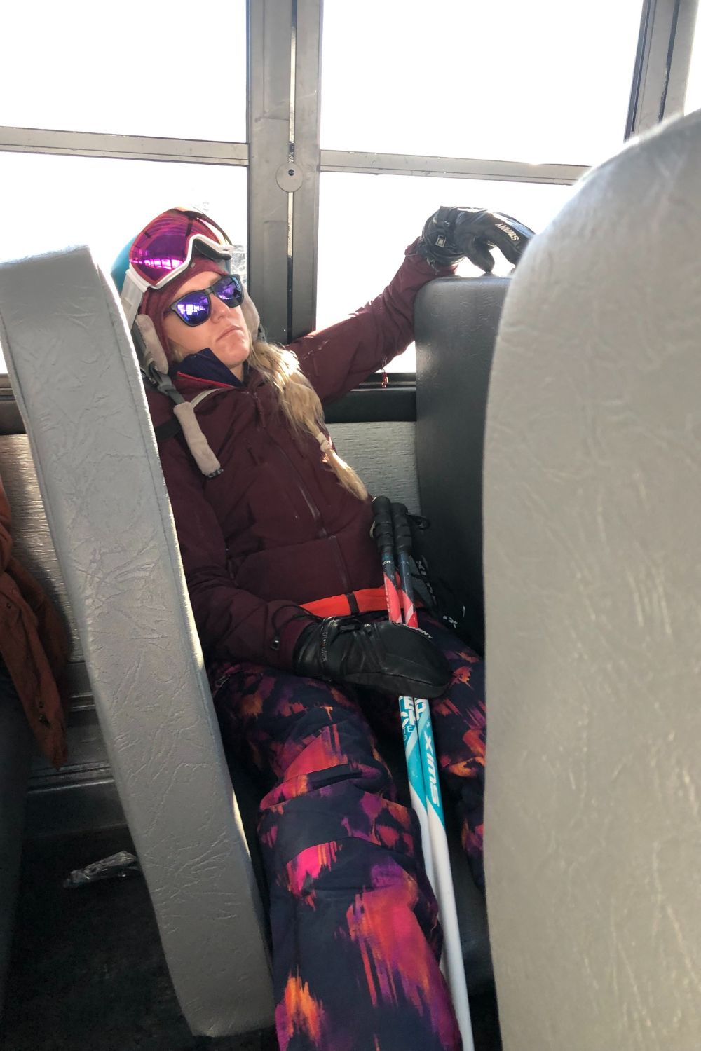 Kate snoozing on a bus wearing her ski gear, including goggles and gloves, with ski poles.