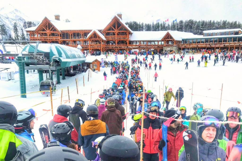 This image shows a large crowd of skiers and snowboarders waiting in line for a ski lift at the base of Lake Louise ski resort. The backdrop features a rustic lodge with snow-covered roofs and vibrant activity on the slopes.