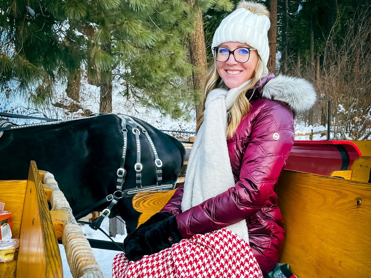 Kate from Kate's Crossing Blog bundled up for winter sits in a horse-drawn sleigh, smiling in a white knit hat, maroon puffer jacket with a fur-trimmed hood, black gloves, and a white scarf. She is draped with a red and white houndstooth blanket. The horse beside her is harnessed and ready for the ride. This cozy winter scene emphasizes the need for warm layers, scarves, and gloves, which are essential items for any "Winter packing list" when traveling to cold, snowy destinations.