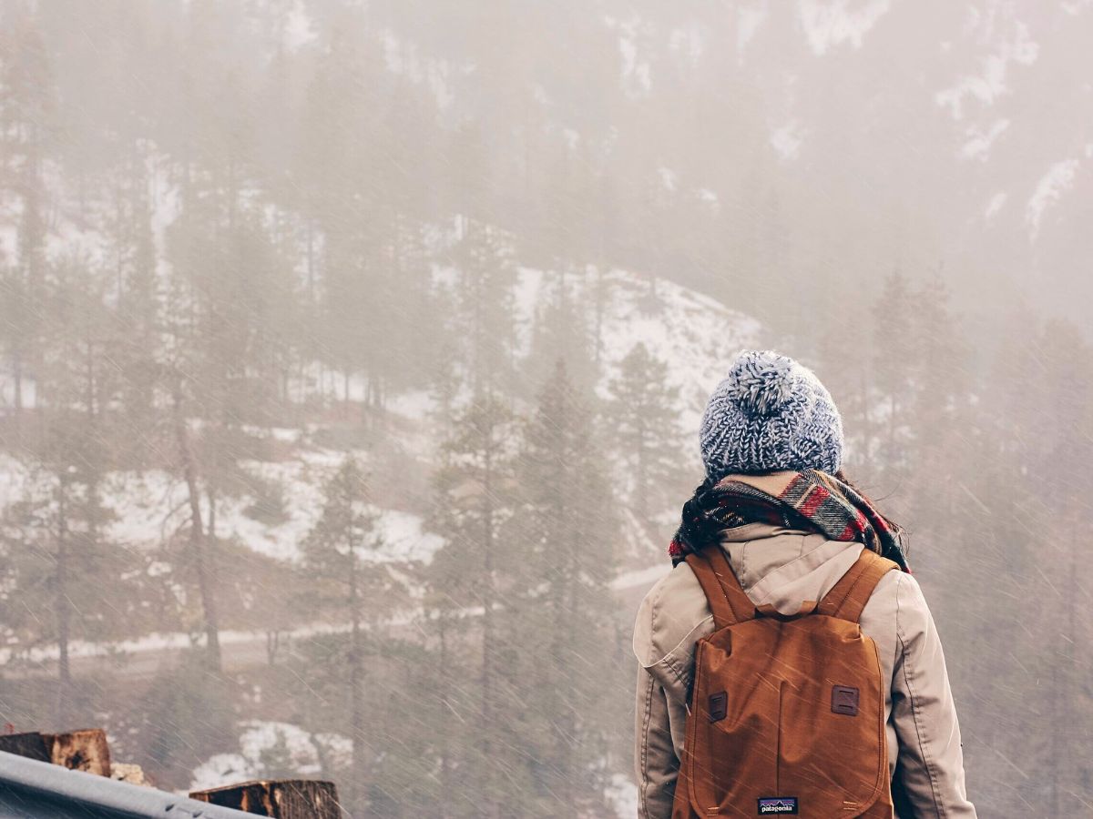 A person stands facing a misty, snow-covered landscape, wearing a cozy knit hat, a plaid scarf, a tan coat, and a brown backpack. The scene captures the quiet beauty of a winter hike through a forested area. This image emphasizes the importance of packing warm, layered clothing, accessories like scarves and hats, and a sturdy backpack for winter explorations.