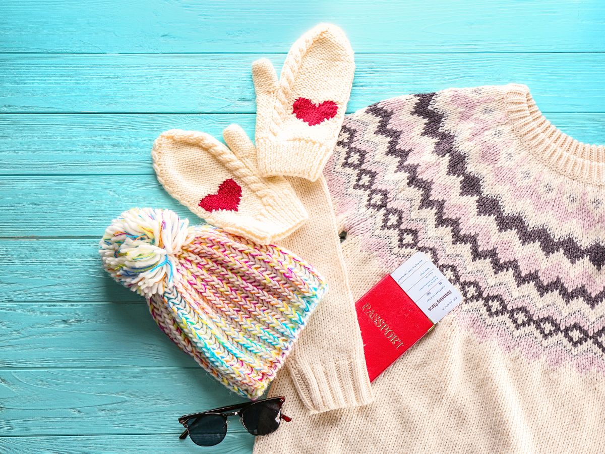 A flat lay image shows cozy winter essentials, including a cream-colored sweater with a geometric pattern, matching knit mittens adorned with red hearts, a multicolored pom-pom hat, sunglasses, and a passport tucked beside the sweater.