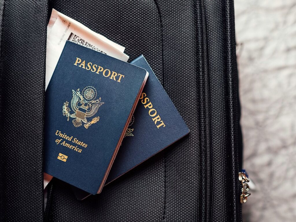 A close-up image shows two U.S. passports tucked into the outer pocket of a black suitcase, with travel documents peeking out from behind them. 