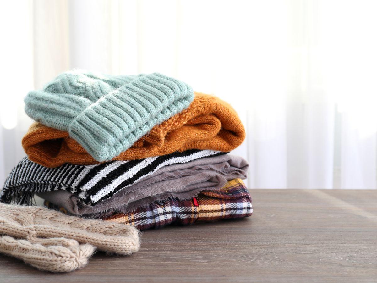 A neatly folded stack of winter clothing sits on a wooden surface, including a light blue knit hat, an orange sweater, a striped scarf, plaid flannel, and a pair of beige knit mittens.