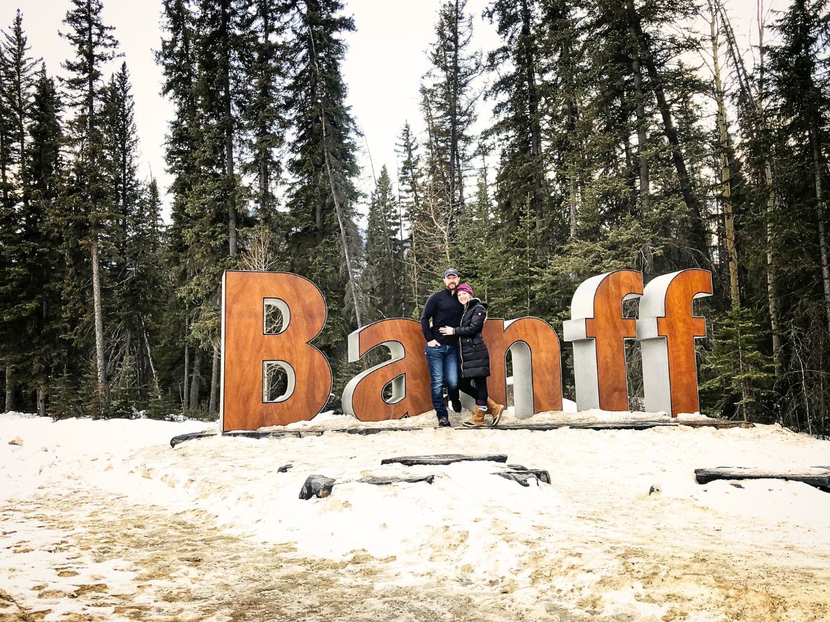 Kate and her husband pose in front of the large "Banff" sign, surrounded by snow-covered ground and tall evergreen trees. Both are dressed warmly in winter jackets, boots, and hats, preparing for the cold weather of the Canadian Rockies.