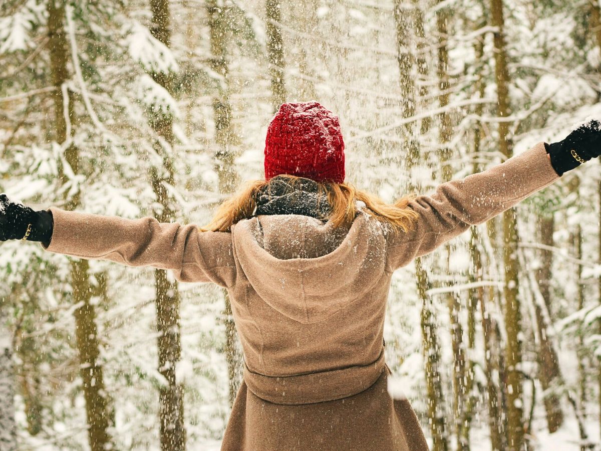 A woman stands in a snow-covered forest with her arms outstretched, enjoying the falling snow. She is wearing a tan winter coat, black gloves, and a red knit hat. This image captures the joy of winter and highlights the need for warm clothing, including coats, hats, and gloves.