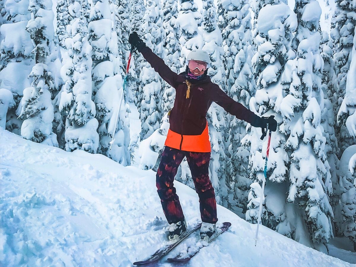 Kate from Kate's Crossing Blog poses with arms raised in excitement on a snowy mountain slope, surrounded by snow-covered trees. Dressed in a maroon jacket, bright orange vest, patterned ski pants, and ski goggles, the skier is well-prepared for the cold weather.