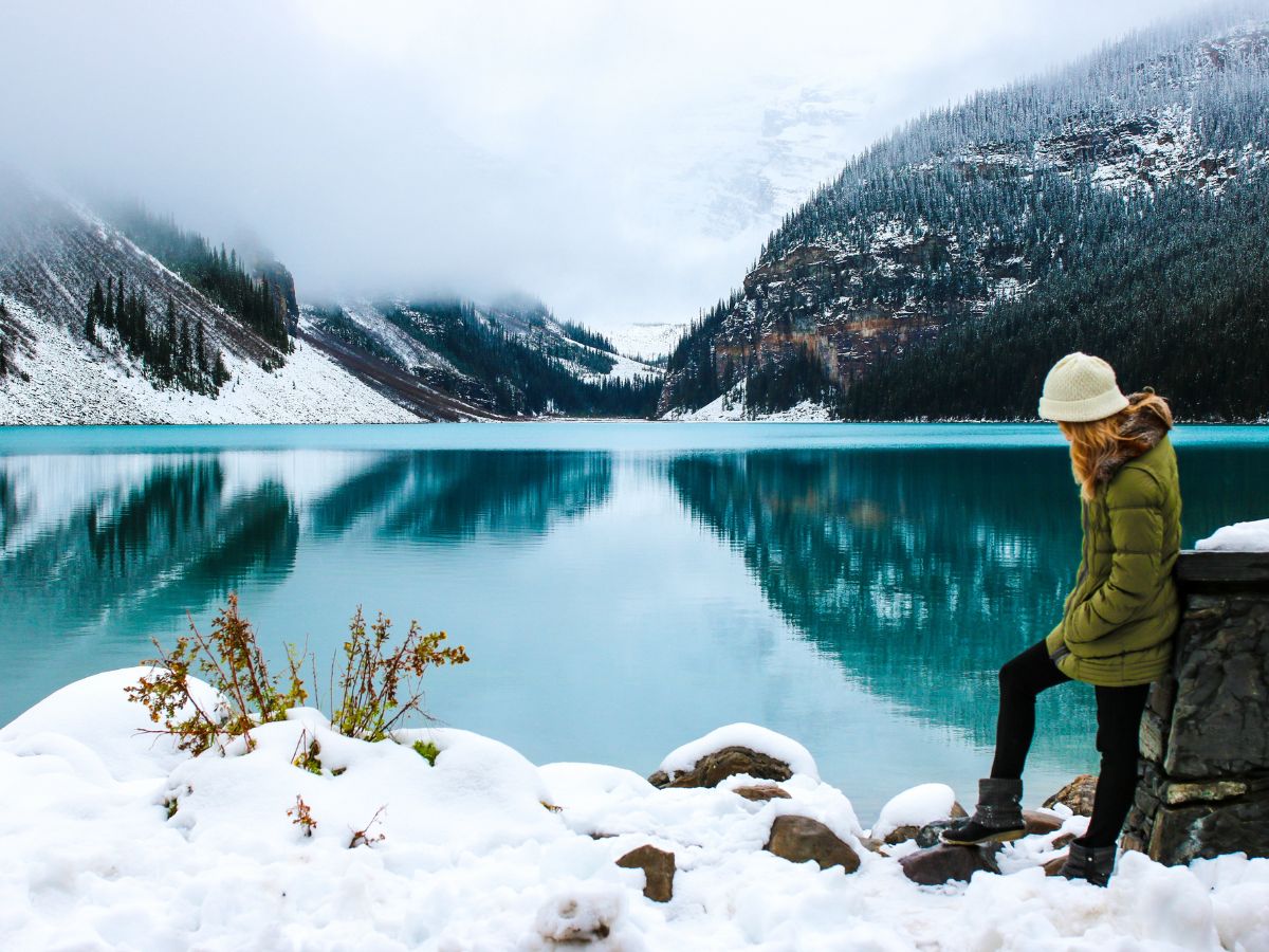 A person dressed warmly in a green winter jacket, black pants, boots, and a white knit hat sits by a snowy lakeside, looking out over a calm, turquoise lake. Snow covers the foreground, and misty mountains rise in the background. The serene winter scene illustrates the importance of dressing warmly for cold-weather adventures, making it perfect for a "Winter packing list" reminder to pack insulated jackets, boots, and hats.