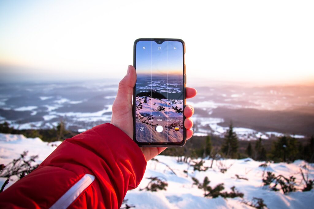 A person holds up a smartphone to capture a snowy mountain view during sunrise or sunset, with the screen displaying the scene as they take a photo. They are wearing a red winter jacket, emphasizing the cold-weather conditions.