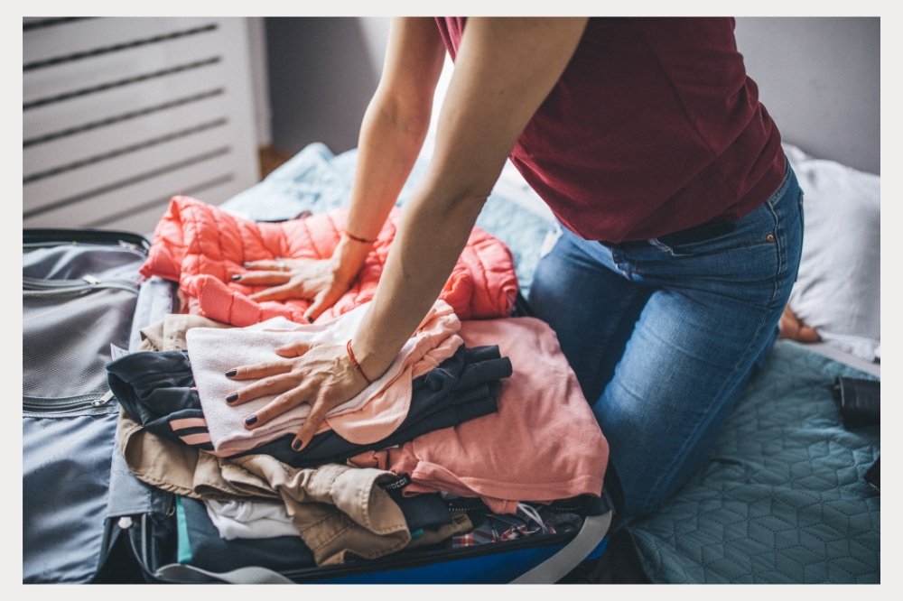 A person is seen packing a suitcase, kneeling on the bed to fit in layers of clothing, including a pink puffer jacket, shirts, and pants. The image captures the last-minute effort to pack efficiently for a trip.