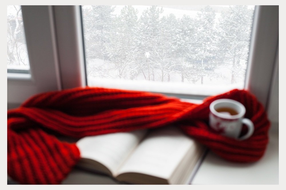 A cozy scene by the window shows a bright red scarf draped over an open book and a mug of tea, with a snowy forest visible outside. This image evokes warmth and relaxation during the winter months, serving as a reminder to pack cozy essentials like scarves and something to enjoy during downtime, like a good book.